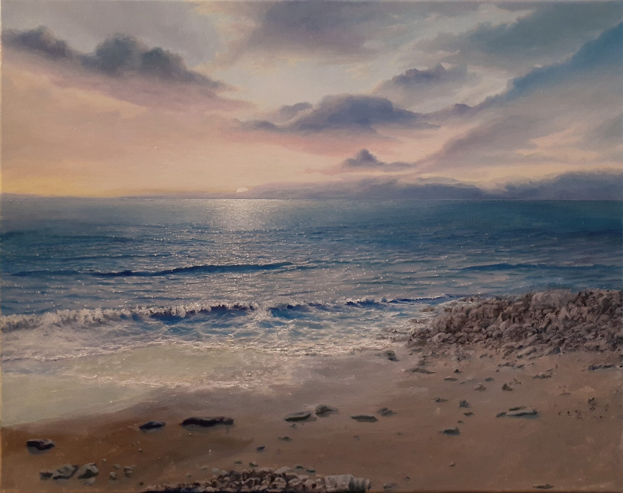 The sun sets over the ocean. There are clouds in the sky and the waves are approaching a pebbled shore.
