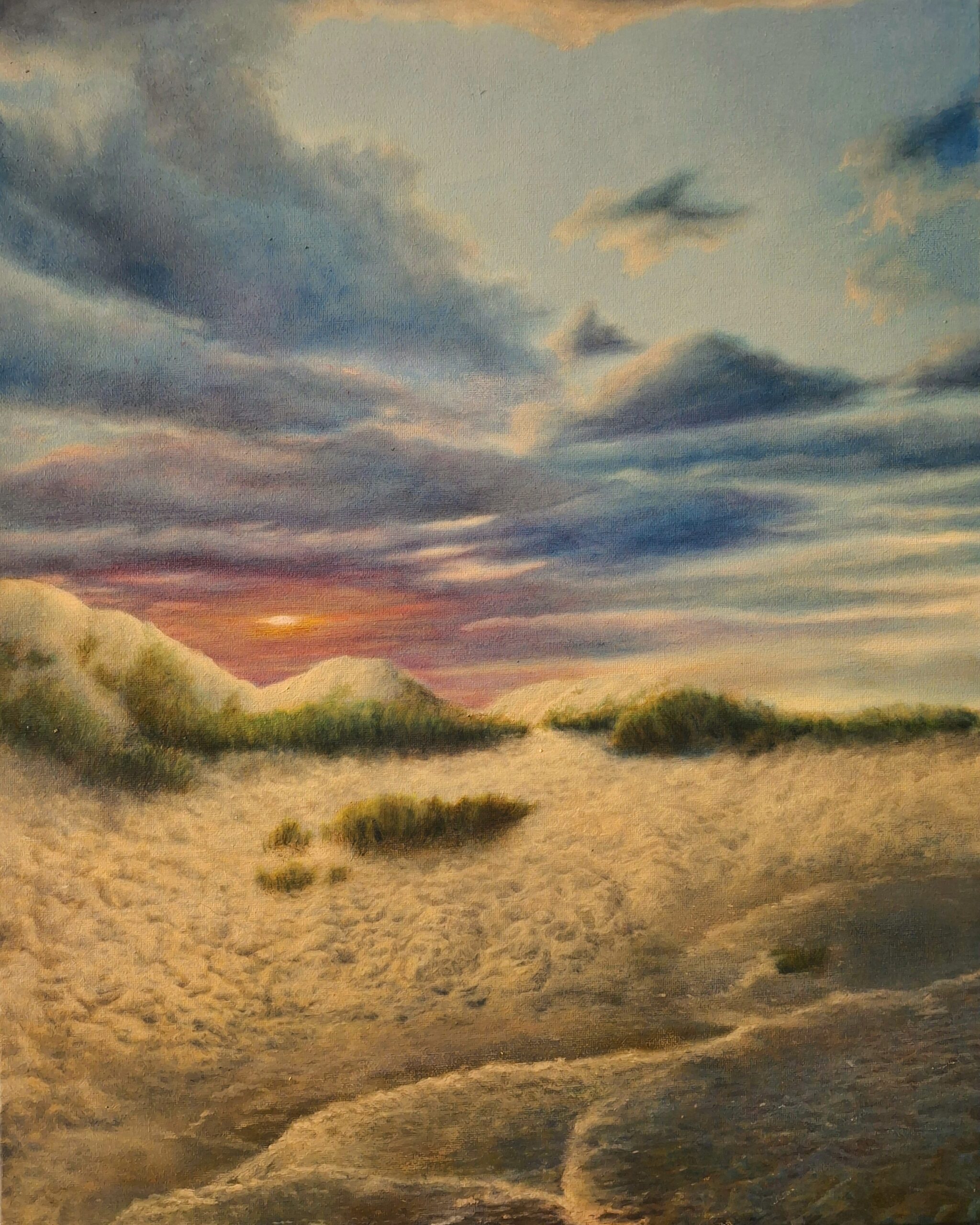 The sun rises over sand dunes. There are coastal grasses in the near distance. In the foreground are a few gentle waves of water.